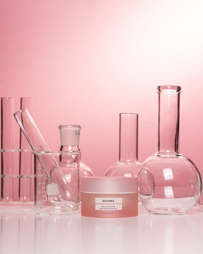 Glass laboratory equipment with a skincare cream against a pink background.